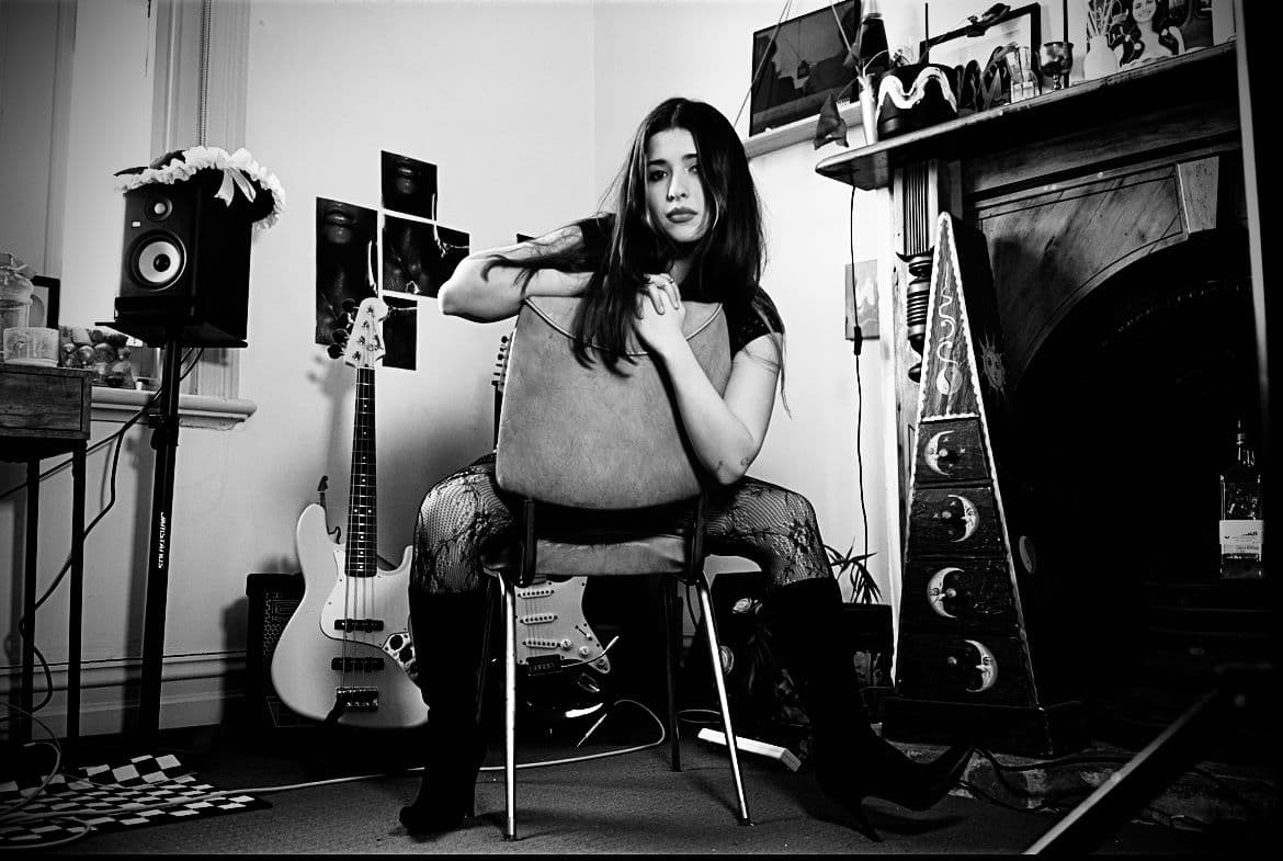 A woman sits in a chair, holding a guitar, looking relaxed and ready to play music.
